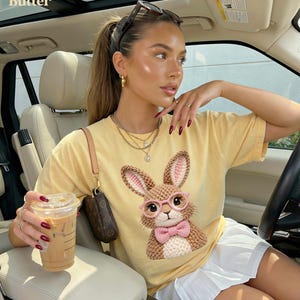 May include: A light yellow t-shirt featuring a detailed illustration of a bunny wearing pink glasses and a bow tie. The bunny has brown and pink crochet details. The word "Butter" is in the top left corner.
