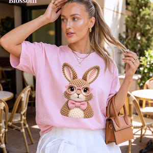 May include: A light pink t-shirt with the word "Blossom" at the top left. The shirt features a detailed graphic of a brown bunny wearing pink glasses and a pink bow tie. The bunny has large ears and is made of a woven material.