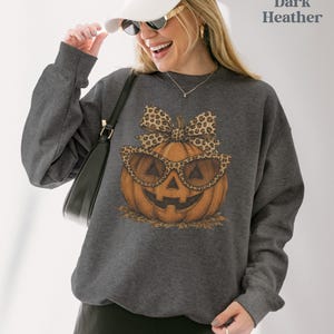 Op de afbeelding: Een donkergrijs gem&ecirc;leerde sweater met ronde hals en een afbeelding van een oranje pompoen met een zonnebril met luipaardprint en een bijpassende strik. De tekst Dark Heather staat in de rechterbovenhoek van de afbeelding.