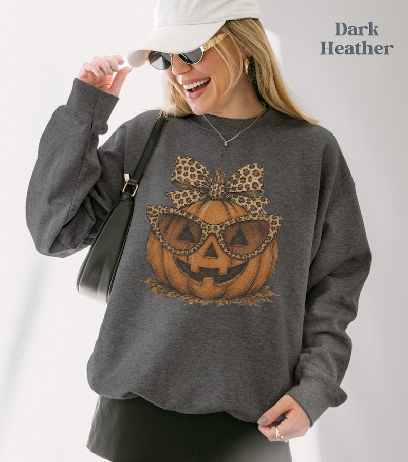 Op de afbeelding: Een donkergrijs gem&ecirc;leerde sweater met ronde hals en een afbeelding van een oranje pompoen met een zonnebril met luipaardprint en een bijpassende strik. De tekst Dark Heather staat in de rechterbovenhoek van de afbeelding.