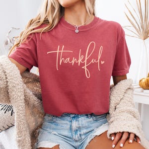 May include: Crimson-colored t-shirt with the word "thankful" in a cursive font. The shirt is paired with denim shorts and a cream-colored cardigan. The image is well-lit, showcasing the casual style.