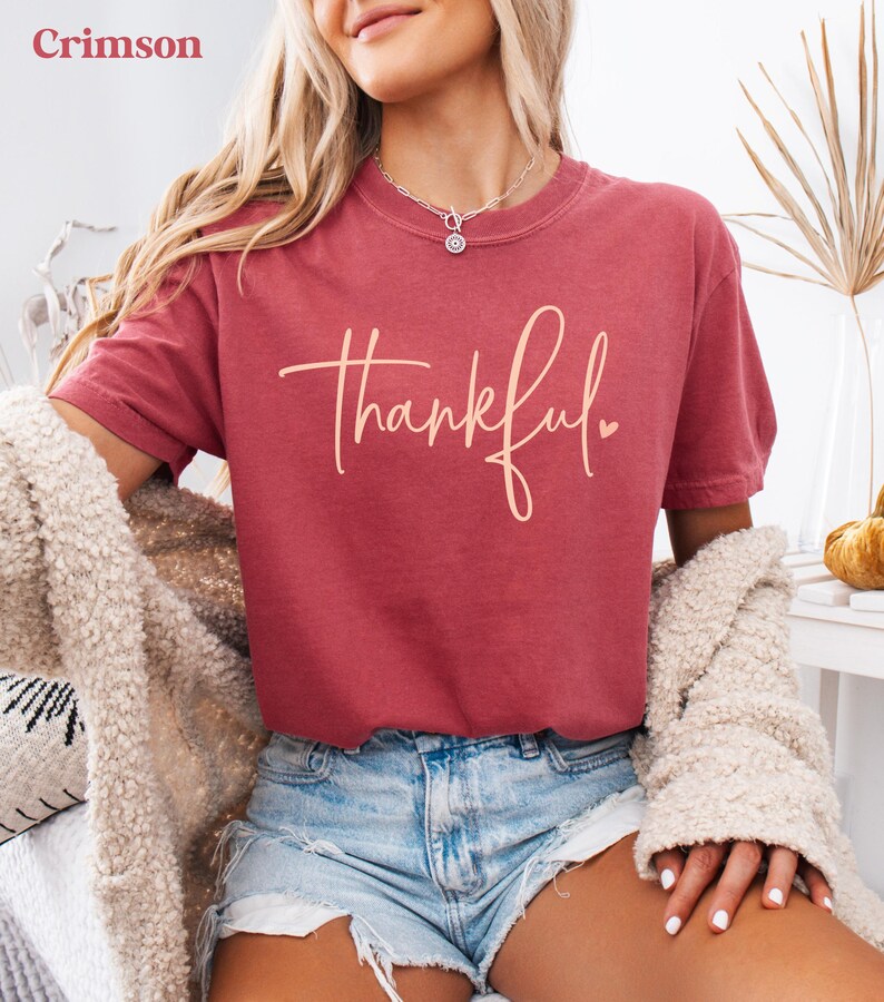 May include: Crimson-colored t-shirt with the word "thankful" in a cursive font. The shirt is paired with denim shorts and a cream-colored cardigan. The image is well-lit, showcasing the casual style.