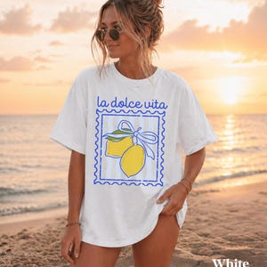 Pu&ograve; includere: T-shirt bianca con la scritta "la dolce vita" sopra una grafica blu con due limoni gialli legati con un fiocco blu. La maglietta ha una vestibilit&agrave; ampia, perfetta per l'uso quotidiano. La parola "White" &egrave; stampata in basso.