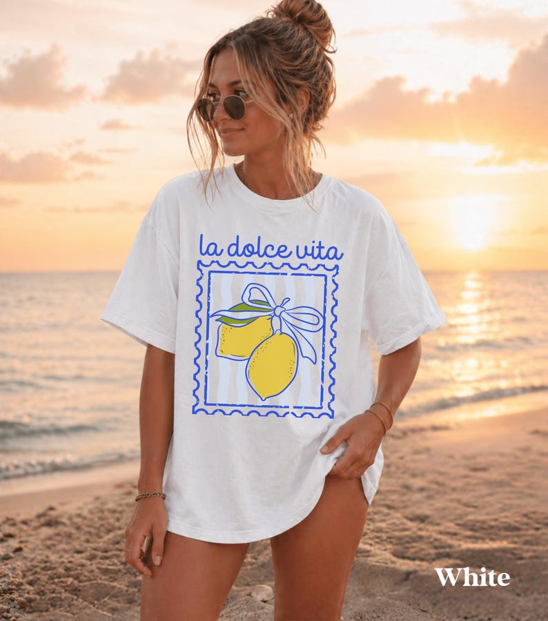 Pu&ograve; includere: T-shirt bianca con la scritta "la dolce vita" sopra una grafica blu con due limoni gialli legati con un fiocco blu. La maglietta ha una vestibilit&agrave; ampia, perfetta per l'uso quotidiano. La parola "White" &egrave; stampata in basso.