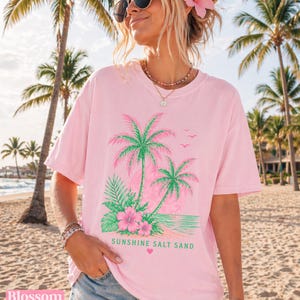 May include: A light pink t-shirt with a tropical design of palm trees, flowers, and the text "Sunshine Salt Sand" in green. The shirt has a relaxed fit and short sleeves. The word "Blossom" is printed at the bottom left.