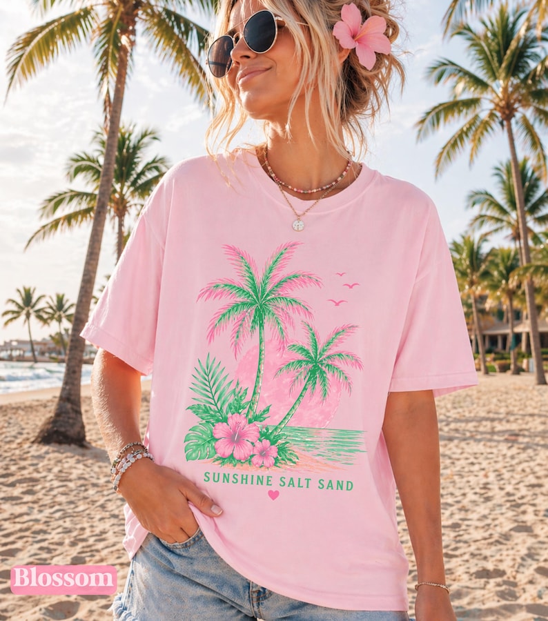 May include: A light pink t-shirt with a tropical design of palm trees, flowers, and the text "Sunshine Salt Sand" in green. The shirt has a relaxed fit and short sleeves. The word "Blossom" is printed at the bottom left.