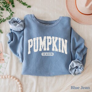 Puede incluir: Una sudadera azul con las palabras "PUMPKIN SEASON" en letras blancas. La sudadera est&aacute; doblada sobre una superficie blanca. Un sombrero es visible en el fondo. Las palabras "Blue Jean" est&aacute;n en la parte inferior.