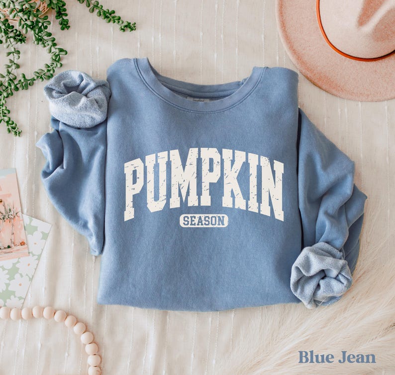 Puede incluir: Una sudadera azul con las palabras "PUMPKIN SEASON" en letras blancas. La sudadera est&aacute; doblada sobre una superficie blanca. Un sombrero es visible en el fondo. Las palabras "Blue Jean" est&aacute;n en la parte inferior.