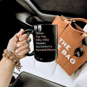 May include: A black ceramic mug with the words "Mama, Tip-Top, Fifty-Fifty, Thanks, Huckleberry, Baseball Player" in white. The mug is held in a hand with gold jewelry. A brown tote bag and sunglasses are in the background.