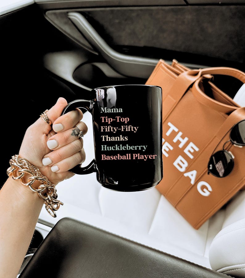 May include: A black ceramic mug with the words "Mama, Tip-Top, Fifty-Fifty, Thanks, Huckleberry, Baseball Player" in white. The mug is held in a hand with gold jewelry. A brown tote bag and sunglasses are in the background.