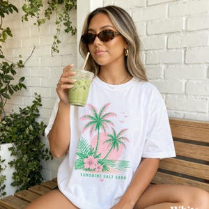 May include: White t-shirt with a tropical design featuring pink palm trees, a pink sun, and the text "Sunshine Salt Sand." The shirt is a relaxed fit and has short sleeves. The model is wearing sunglasses and holding a green drink.