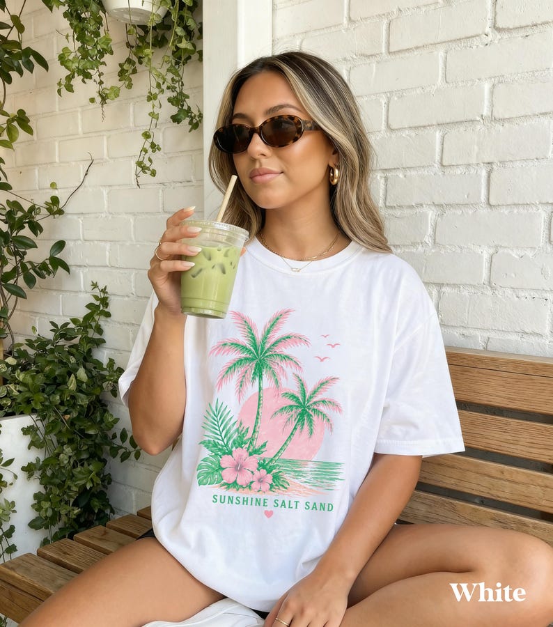 May include: White t-shirt with a tropical design featuring pink palm trees, a pink sun, and the text "Sunshine Salt Sand." The shirt is a relaxed fit and has short sleeves. The model is wearing sunglasses and holding a green drink.