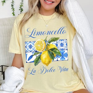 Pu&ograve; includere: Una maglietta gialla con una grafica di limoni, foglie e piastrelle blu e bianche. Il testo "Limoncello La Dolce Vita" &egrave; stampato sulla maglietta.
