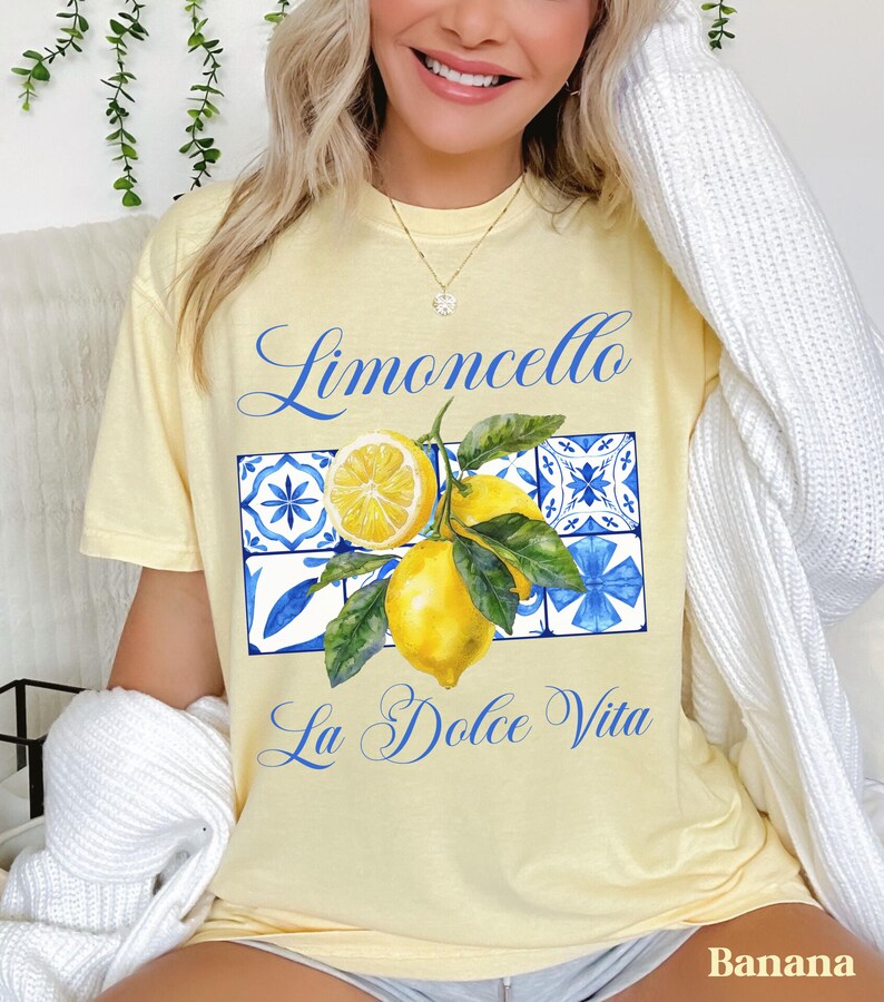 Pu&ograve; includere: Una maglietta gialla con una grafica di limoni, foglie e piastrelle blu e bianche. Il testo "Limoncello La Dolce Vita" &egrave; stampato sulla maglietta.