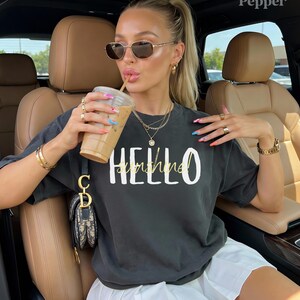 May include: Dark grey t-shirt with the words "HELLO sunshine" in white and gold. The person is wearing sunglasses, gold jewellery, and holding a coffee drink. The person is sitting in a car.