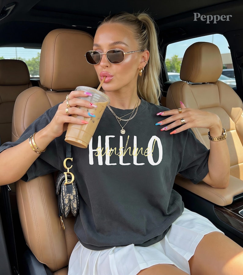 May include: Dark grey t-shirt with the words "HELLO sunshine" in white and gold. The person is wearing sunglasses, gold jewellery, and holding a coffee drink. The person is sitting in a car.