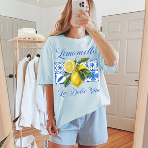 Pu&ograve; includere: Una maglietta azzurra con una grafica di limoni e piastrelle blu e bianche. Il testo "Limoncello La Dolce Vita" &egrave; stampato sulla maglietta.