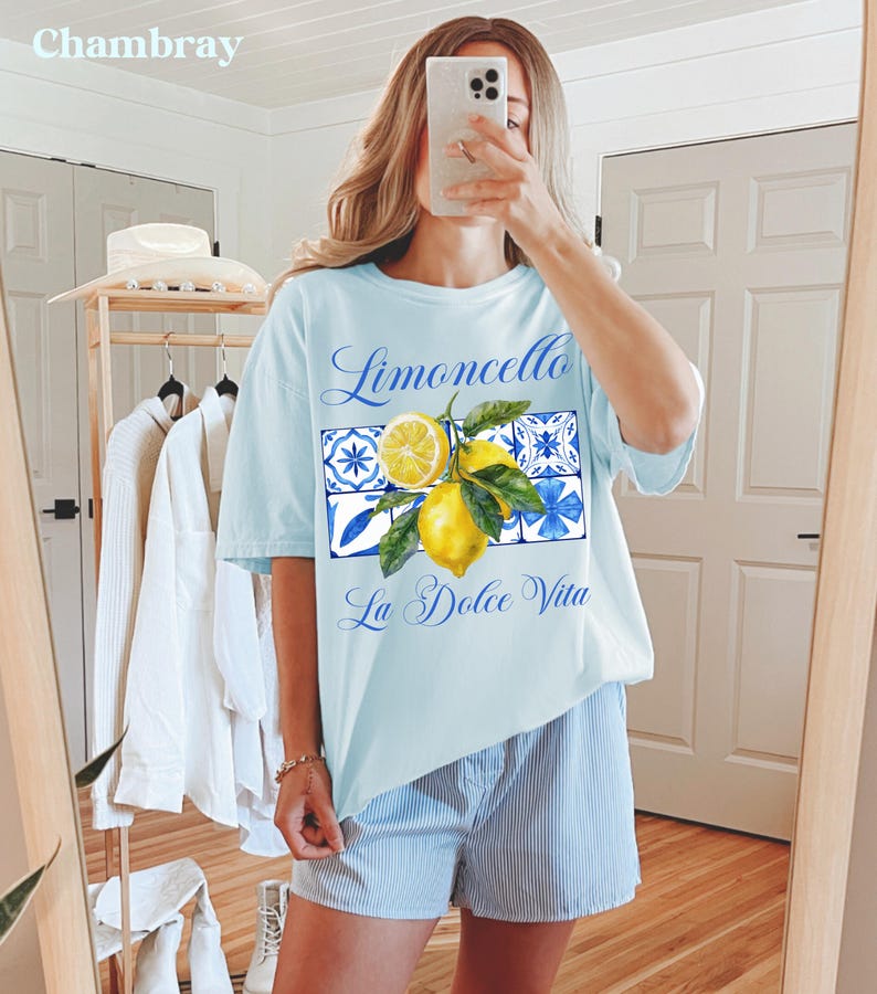 Pu&ograve; includere: Una maglietta azzurra con una grafica di limoni e piastrelle blu e bianche. Il testo "Limoncello La Dolce Vita" &egrave; stampato sulla maglietta.