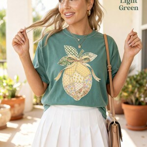 May include: Light green t-shirt featuring a lemon graphic. The lemon design is a patchwork of different patterns, including gingham and floral prints. The text "Light Green" is in the upper right corner. The model is wearing a white pleated skirt and a brown handbag.