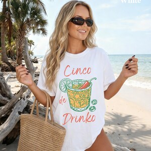 May include: White t-shirt with a Cinco de Drinko graphic. The design features a cocktail with a lime, chili pepper, and ice cubes. The shirt is worn on a beach with a straw tote bag.