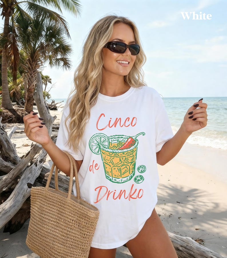 May include: White t-shirt with a Cinco de Drinko graphic. The design features a cocktail with a lime, chili pepper, and ice cubes. The shirt is worn on a beach with a straw tote bag.