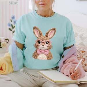 May include: A light blue t-shirt featuring a crocheted bunny wearing pink glasses and a bow tie. The bunny has brown fur and pink inner ears. The text "Chalky Mint" is in the upper left corner.