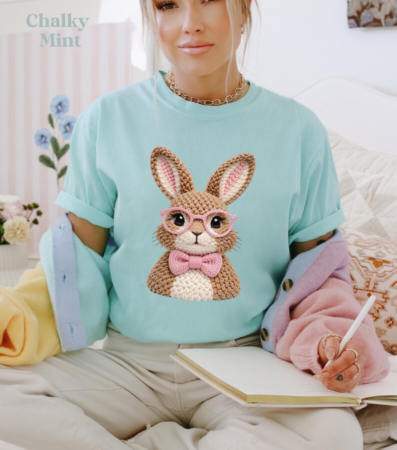 May include: A light blue t-shirt featuring a crocheted bunny wearing pink glasses and a bow tie. The bunny has brown fur and pink inner ears. The text "Chalky Mint" is in the upper left corner.
