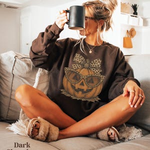 Op de afbeelding: Een donkerchocoladebruin sweatshirt met lange mouwen en een centrale afbeelding van een oranje pompoen met een bril met luipaardprint en een bijpassende strik. Het sweatshirt is effen donkerbruin met een ronde hals en geribbelde boorden.