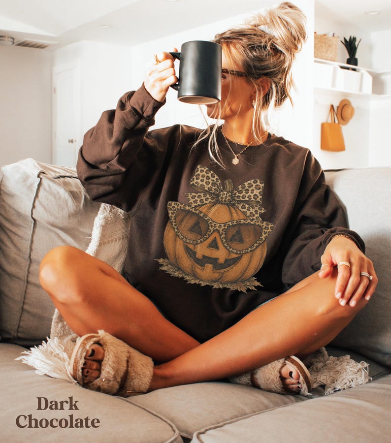 Op de afbeelding: Een donkerchocoladebruin sweatshirt met lange mouwen en een centrale afbeelding van een oranje pompoen met een bril met luipaardprint en een bijpassende strik. Het sweatshirt is effen donkerbruin met een ronde hals en geribbelde boorden.