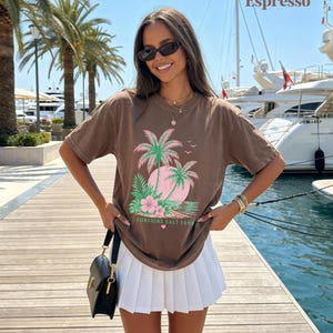 May include: Espresso-coloured t-shirt with a tropical design of palm trees, a pink sun, and the text "Sunshine Salt Sand." The shirt is worn with a white pleated skirt and a black handbag. The background shows palm trees and boats.