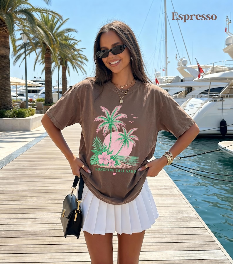May include: Espresso-coloured t-shirt with a tropical design of palm trees, a pink sun, and the text "Sunshine Salt Sand." The shirt is worn with a white pleated skirt and a black handbag. The background shows palm trees and boats.