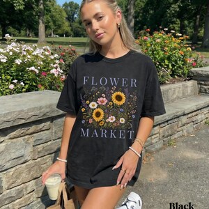 May include: A black short-sleeve t-shirt featuring a graphic print of a colorful wildflower bouquet with sunflowers, daisies, and lavender. The text FLOWER MARKET is printed in a light purple serif font above and below the floral illustration.