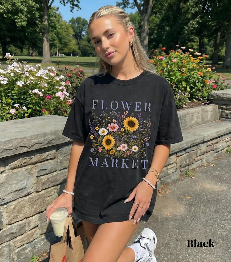 May include: A black short-sleeve t-shirt featuring a graphic print of a colorful wildflower bouquet with sunflowers, daisies, and lavender. The text FLOWER MARKET is printed in a light purple serif font above and below the floral illustration.