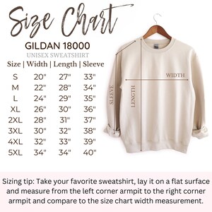 May include: A beige Gildan 18000 unisex sweatshirt displayed on a wooden hanger with an accompanying size chart. The chart lists measurements in inches for sizes S through 5XL, detailing width, length, and sleeve dimensions for the garment.
