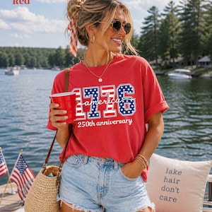 May include: A red t-shirt with the text "1776 America 250th anniversary". The design incorporates the American flag's colours and stars. The shirt's text includes the numbers 1776 in a blue and white stars and stripes pattern, with the words "America" and "250th anniversary" in white.