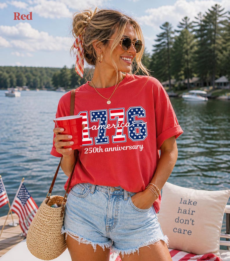May include: A red t-shirt with the text "1776 America 250th anniversary". The design incorporates the American flag's colours and stars. The shirt's text includes the numbers 1776 in a blue and white stars and stripes pattern, with the words "America" and "250th anniversary" in white.