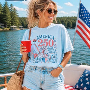 K&ouml;nnte beinhalten: Hellblaues T-Shirt mit dem Text "AMERICA 250th Sweet Land of Liberty 1776-2026" und einer Grafik der Freiheitsstatue. Das Shirt wird mit Jeansshorts kombiniert. Im Hintergrund ist eine amerikanische Flagge zu sehen.