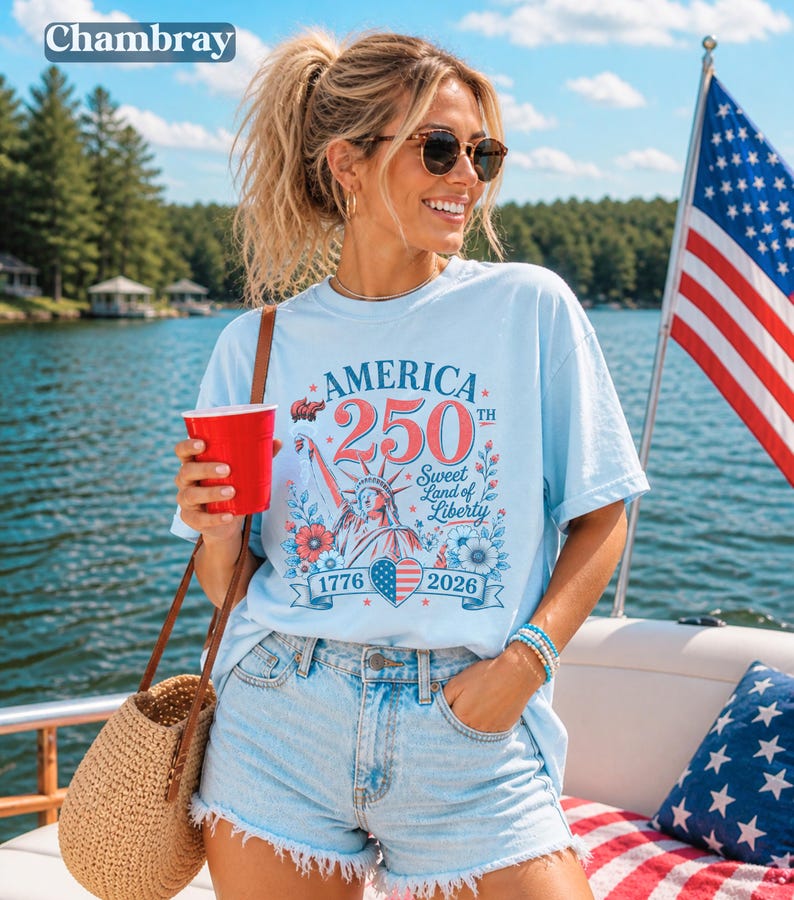 K&ouml;nnte beinhalten: Hellblaues T-Shirt mit dem Text "AMERICA 250th Sweet Land of Liberty 1776-2026" und einer Grafik der Freiheitsstatue. Das Shirt wird mit Jeansshorts kombiniert. Im Hintergrund ist eine amerikanische Flagge zu sehen.