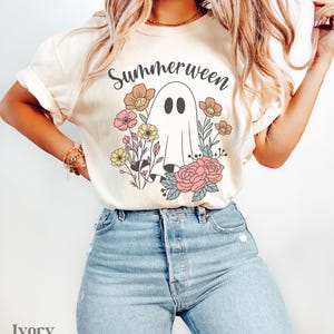May include: Ivory-colored t-shirt with the word "Summerween" above a ghost illustration surrounded by flowers in pink, yellow, and orange. The model is wearing light blue jeans.
