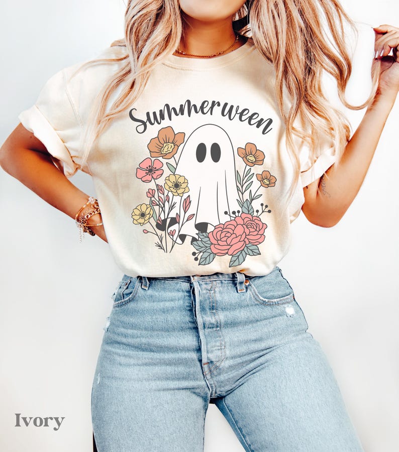 May include: Ivory-colored t-shirt with the word "Summerween" above a ghost illustration surrounded by flowers in pink, yellow, and orange. The model is wearing light blue jeans.