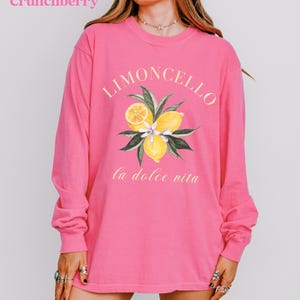 May include: A long-sleeved, bright pink shirt with the word "Limoncello" in a yellow script above a watercolor illustration of lemons and leaves. The phrase "la dolce vita" is printed below the illustration. The word "Crunchberry" is in the top left corner.