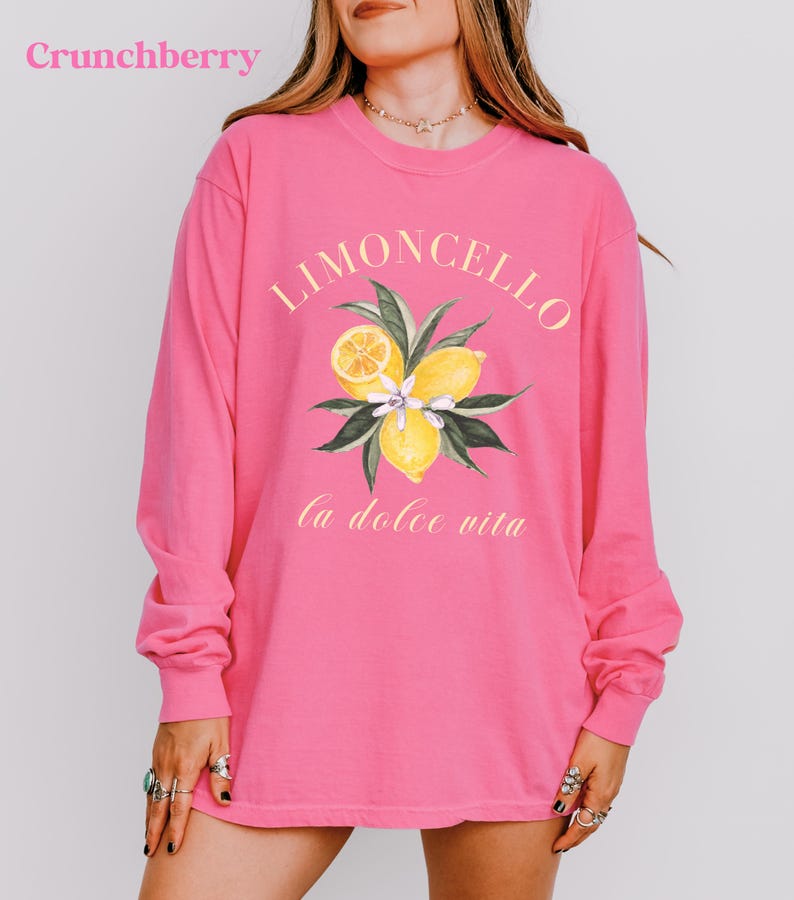 May include: A long-sleeved, bright pink shirt with the word "Limoncello" in a yellow script above a watercolor illustration of lemons and leaves. The phrase "la dolce vita" is printed below the illustration. The word "Crunchberry" is in the top left corner.