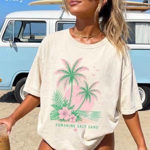 May include: Ivory t-shirt with a tropical design featuring palm trees, pink flowers, and the text "Sunshine Salt Sand." The graphic is printed on a relaxed-fit tee. A light blue van is in the background.