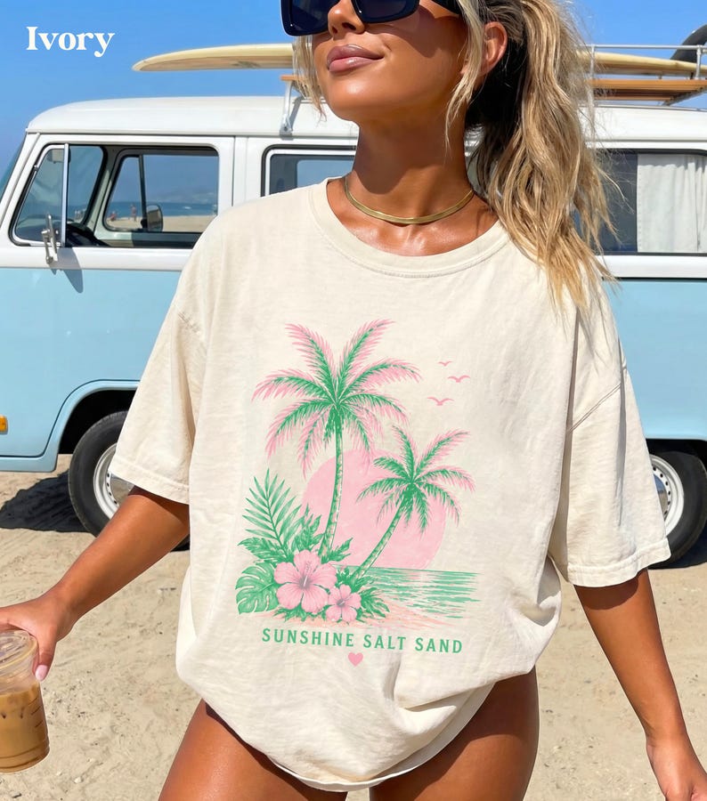 May include: Ivory t-shirt with a tropical design featuring palm trees, pink flowers, and the text "Sunshine Salt Sand." The graphic is printed on a relaxed-fit tee. A light blue van is in the background.