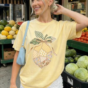 May include: A pale yellow t-shirt with a lemon design made of patchwork fabric. The lemon has a green leaf and bow, with floral, gingham, and polka dot patterns. The word "Butter" is printed at the bottom of the image.