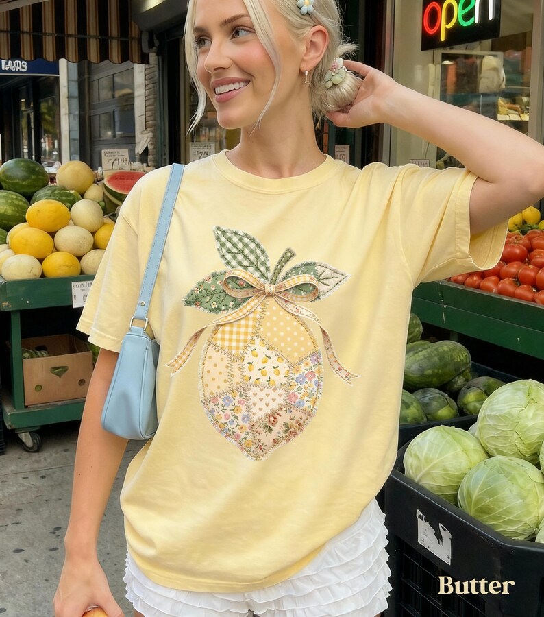 May include: A pale yellow t-shirt with a lemon design made of patchwork fabric. The lemon has a green leaf and bow, with floral, gingham, and polka dot patterns. The word "Butter" is printed at the bottom of the image.
