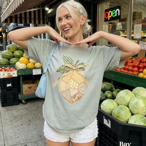 May include: A pale green t-shirt with a patchwork lemon design and a bow. The outfit includes white ruffled shorts and a light blue shoulder bag. The word "Bay" is visible in the bottom right corner.