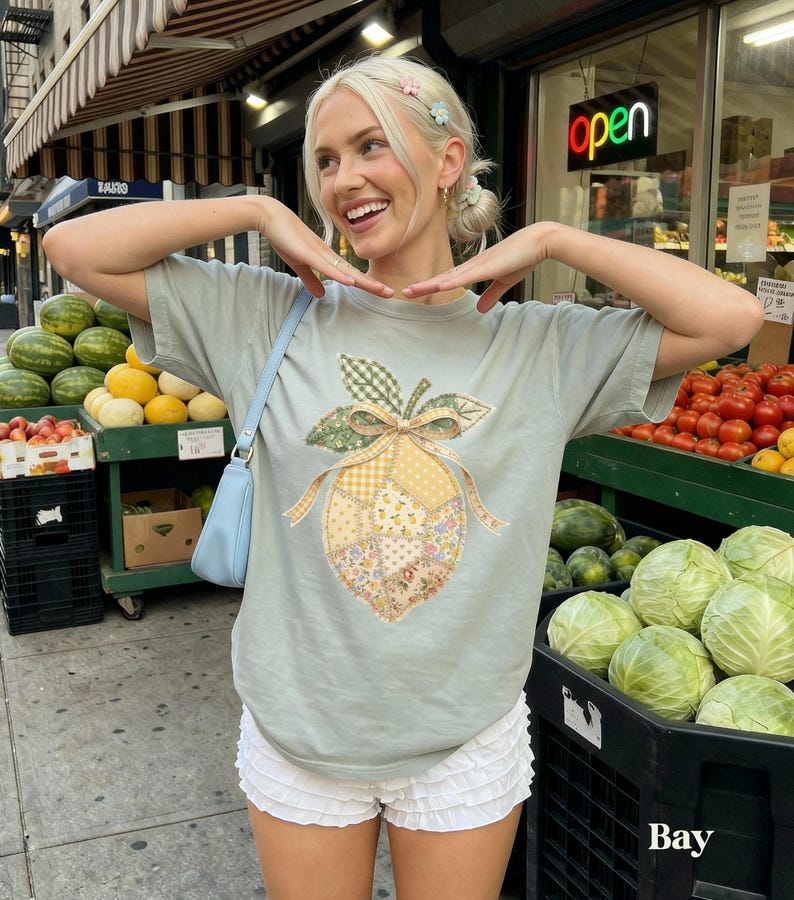 May include: A pale green t-shirt with a patchwork lemon design and a bow. The outfit includes white ruffled shorts and a light blue shoulder bag. The word "Bay" is visible in the bottom right corner.