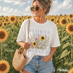 May include: A white short-sleeved t-shirt featuring a graphic print of various wildflowers and sunflowers with the text FLOWER MARKET in light purple lettering, paired with light blue denim shorts and a woven straw handbag with round handles.