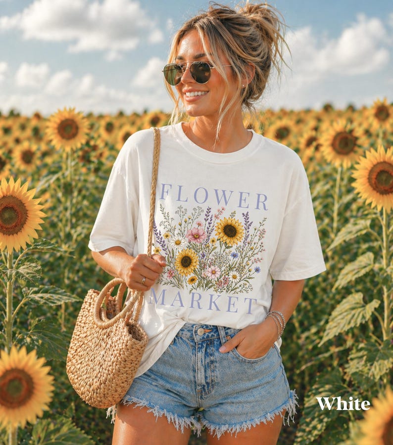 May include: A white short-sleeved t-shirt featuring a graphic print of various wildflowers and sunflowers with the text FLOWER MARKET in light purple lettering, paired with light blue denim shorts and a woven straw handbag with round handles.